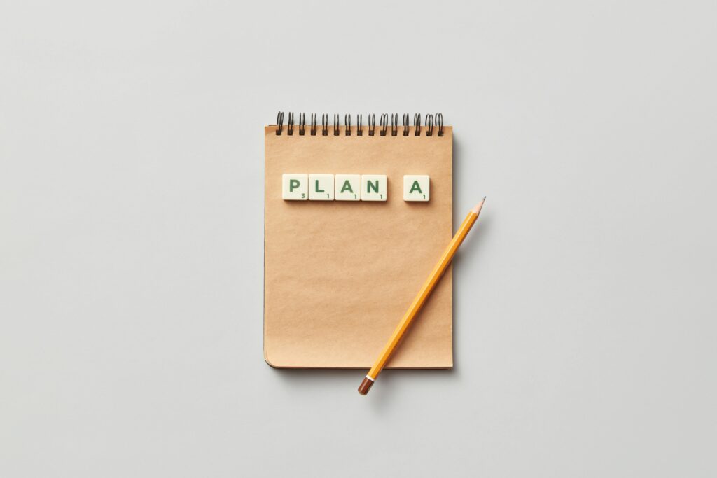 Top view of a notepad with a pencil and scrabble tiles spelling 'PLAN A' on a gray background.